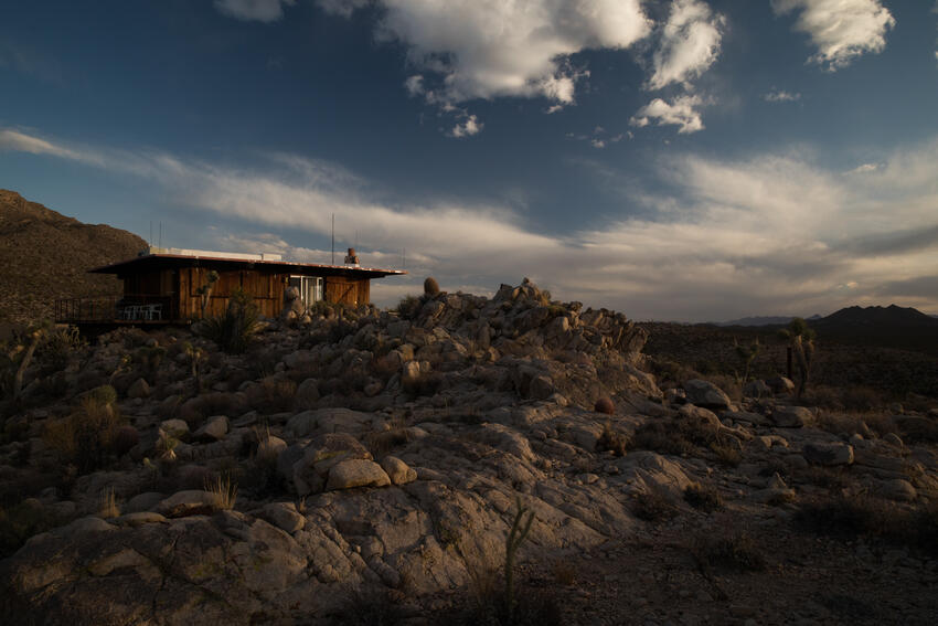 A photo of a wide desert landscape, with mountains on the horizon.  A small wooden house sits in the foreground.