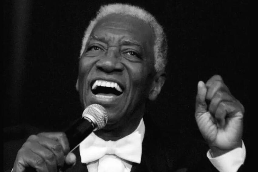 Joe Williams, dressed in a black tux with a white tie, sings into a microphone while gesticulating.