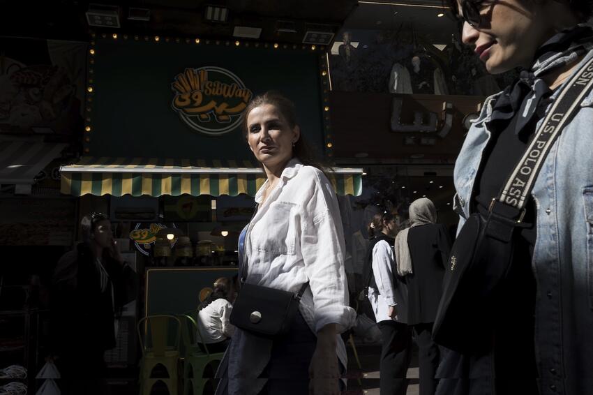 Well-dressed women walk along a city street past stores selling food and clothing. The sign behind them is written in English and Farsi. One woman in the background covers her hair with a headscarf; the rest are bareheaded.