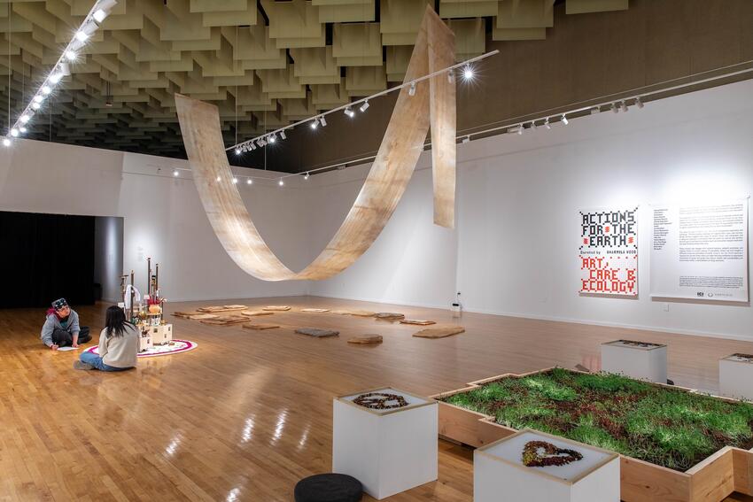 An open interior with a wooden floor. A banner reading, “Actions for the Earth: Art, Care &amp; Ecology” hangs on the wall. A mottled brown strip of fabric hangs from the high ceiling like a giant hammock. Flat brown cushions lie on the floor under the arc of cloth. A wooden box in the foreground holds brown soil and green plants. Dried beans have been arranged into pictures on pedestals around the planter box. Two people are sitting on the floor near a detailed sculpture off to the side.