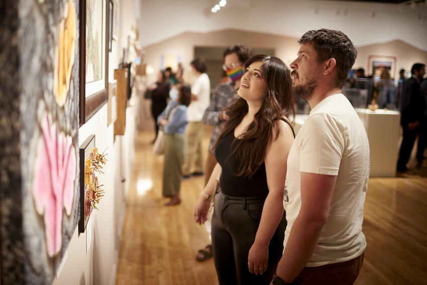 A man and a woman in profile, viewing several works of art ranging from mixed media, photography and a vibrant, abstract painting of a Cholla Cactus featured in the Spirit of the Land exhibition, during the Spring 2022 opening reception.