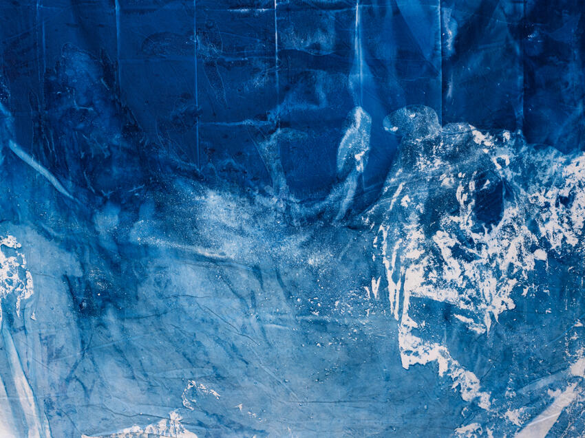 A constellation of white-blue flecks and pale blue washes flows like a breaking wave across a deep blue surface. A rosette of white, crumpled marks breaks into the blue on the right side of the image. Vertical pale lines run like creases across the top, as if the surface has been folded.