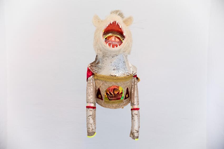 A sculpture hovers in a white space. It resembles a torso with long arms and a hairy catlike head. The red fanged mouth is open. The neck and arms are made of gleaming silver sequins. The torso is made from woven strips of pale fiber with an iridescent logo on the front. The logo looks like a stylized snake in a basket.
