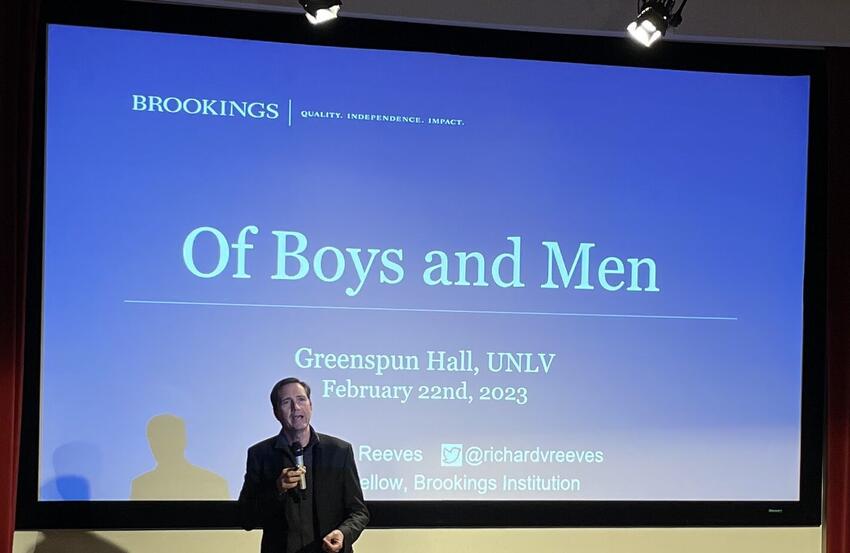 Richard V. Reeves delivering his "Of Boys and Men" lecture on February 22. He stands before a large screen displaying the name of his lecture on a blue PowerPoint background.