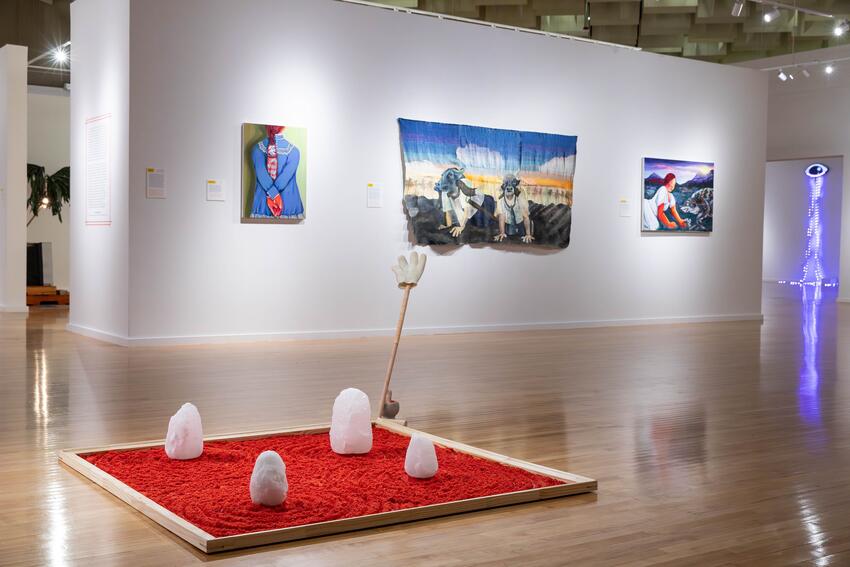 Three artworks hang on a white wall in a gallery. The one in the middle is the largest—a picture of two people with buffalo heads charging over a ploughed field on all fours. The one to the left depicts a woman with her back to us; the one on the right shows us a woman facing a coyote. A zen garden made from red sand-like material lies on the wooden floor in front of the wall. Four white stones stand upright in the square of red. A rake is positioned upright in one corner of the square.