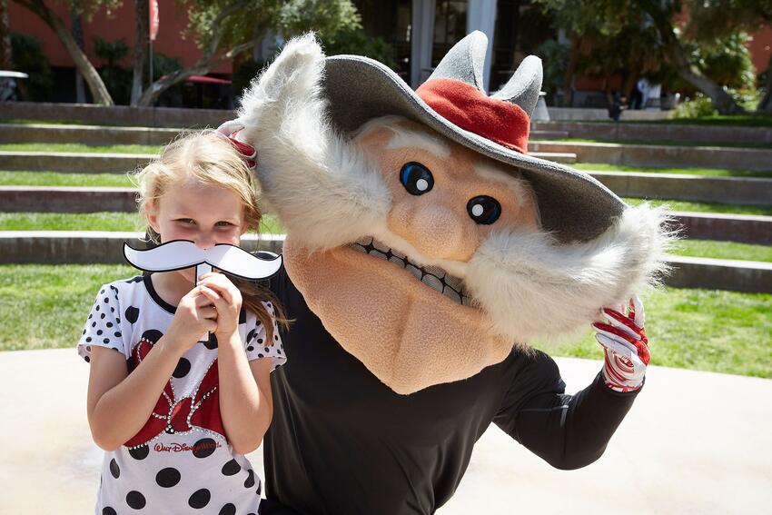 Child holding a U-N-L-V mustache posing with Hey Reb!