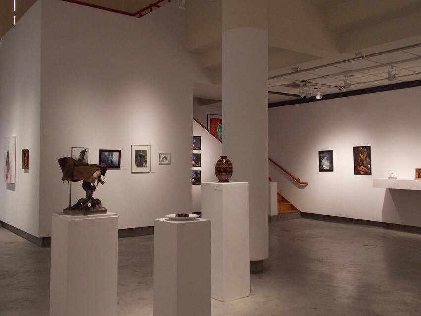 A gallery with multiple pieces of framed art work on the walls and sculptures sitting on pedestals