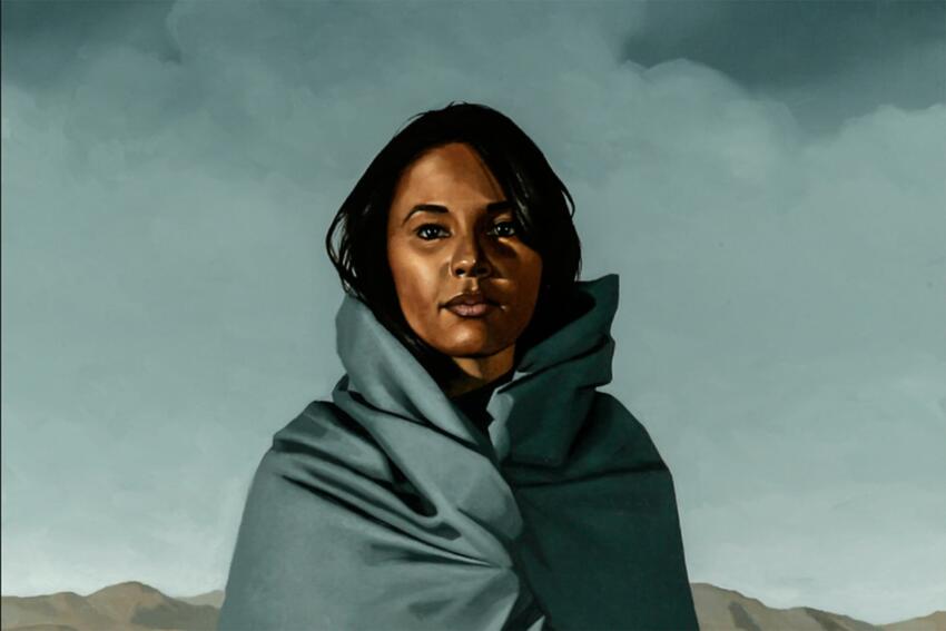 Artwork of a woman wrapped in a colorful robe on a desert landscape with mountains in the distance behind her.
