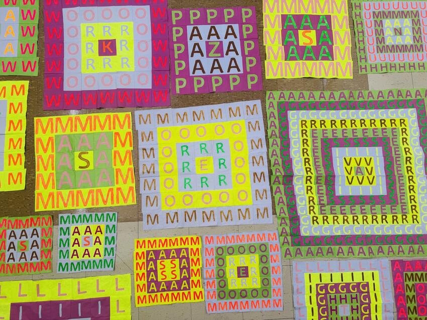 Bright cloth squares lie on the floor. Each square is decorated with a grid. Each square in each grid contains a letter. The squares and the letters are painted with vivid, contrasting colors -- green, yellow, pink, orange, purple.
