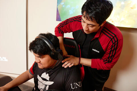 A man adjusts the shoulder of another man playing a computer game.