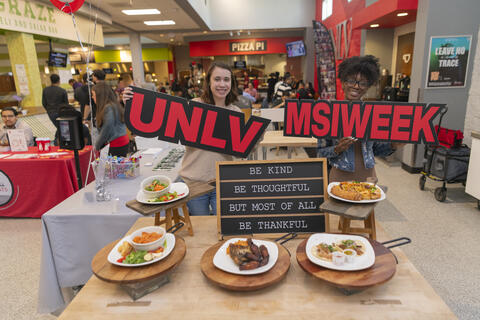 two students in a dining area holding signs that say "UNLV" and "MSI WEEK"