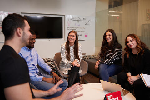 Five people sit in a small meeting area, smiling and talking together with a whiteboard and screen visible in the background.