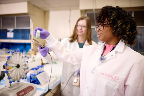 Two people wearing lab coats and gloves work in a laboratory, with one holding a small sample container.