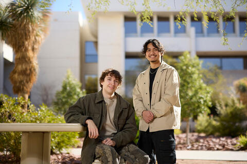 Two students pose outdoors, one sitting down at a table and the other standing besides him.